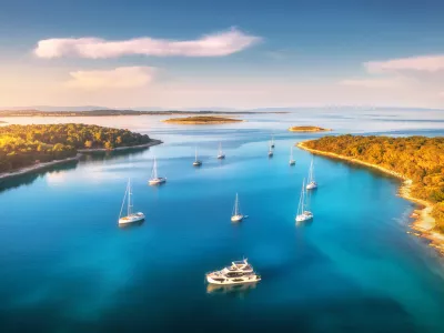 Aerial view of beautiful yachts and boats on the sea at sunset in summer. Adriatic sea, Kamenjak, Croatia. Top view of luxury yachts, sailboats, lagoon, clear blue water, and green forest. Travel