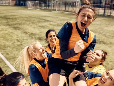 Cheerful team of female soccer players celebrating victory and carrying on of teammates who is shouting out of joy on stadium.