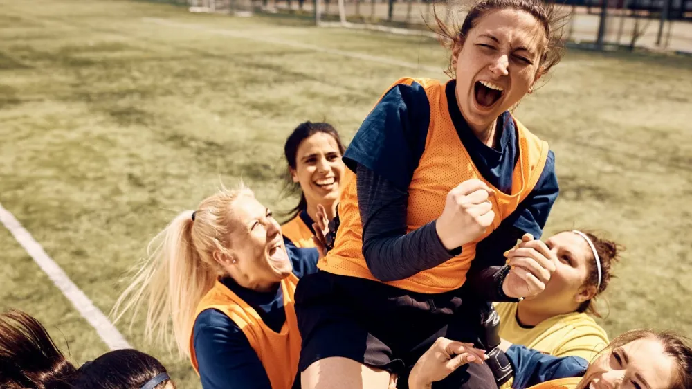 Cheerful team of female soccer players celebrating victory and carrying on of teammates who is shouting out of joy on stadium.
