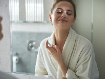 Self care concept. Waist up reflection of young smiling woman in white bathrobe enjoying her morning while applying favorite perfume before bathroom mirror