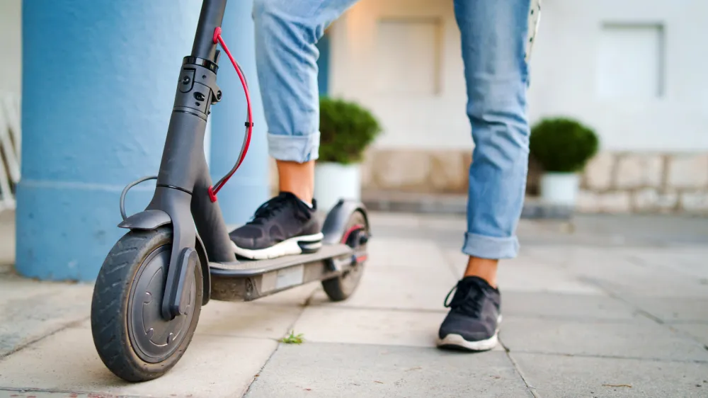 Close up on woman legs feet standing on the electric kick scooter on the pavement wearing jeans and sneakers in summer day