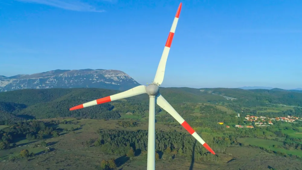 AERIAL CLOSE UP: Spring sunrays illuminating white metal blades in green countryside. Wind-turbine generator spinning in strong winds, representing a renewable source of electricity for village nearby
