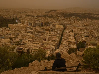 A tourist sits on a bench at the Lycabettus hill as the city of Athens with the ancient Acropolis hill is seen at the background, on Tuesday, April 23, 2024. The Acropolis and other Athens landmarks took on Martian hues Tuesday as stifling dust clouds blown across the Mediterranean Sea from North Africa engulfed the Greek capital. (AP Photo/Petros Giannakouris)