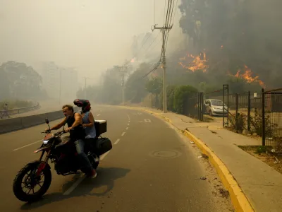 Residents evacuate on a motorcycle as smoke caused by forest fires fill the sky and flames spread into Vina del Mar, Chile, Saturday, Feb. 3, 2024. Officials say intense forest fires burning around a densely populated area of central Chile have left several people dead and destroyed hundreds of homes. (AP Photo/Esteban Felix)