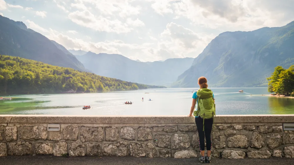 Young woman wearing green backpack is standing on the bridge near Bohinj lake looking at the view in Bohinj, Slovenia
