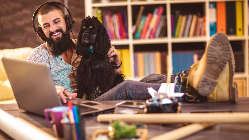 Handsome tattooed man working at home on laptop while sitting at the table with cute dog