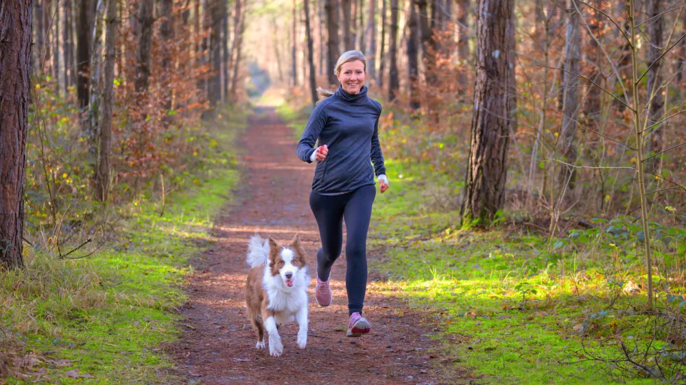 Healthy fit woman running with her dog jogging together through a forest along a footpath approaching the camera with a happy smile / Foto: Mheim3011