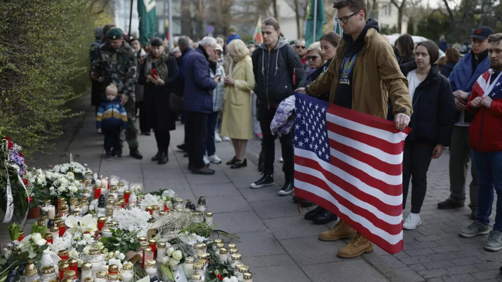 A man with a U.S. national flag and other people stand as they come to pay their last respects in memory of three U.S. Army soldiers found dead on Monday at a training range in Pabrade, near the U.S. Embassy in Vilnius, Lithuania, Tuesday, April 1, 2025. (AP Photo/Mindaugas Kulbis)
