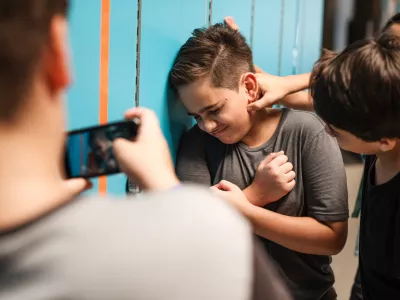 A young boy is being bullied by peers at school. The boy is upset as another boy grabs his neck. A third boy records the bullying incident on his phone. Bullying of teenager in high scool. / Foto: Rawpixel