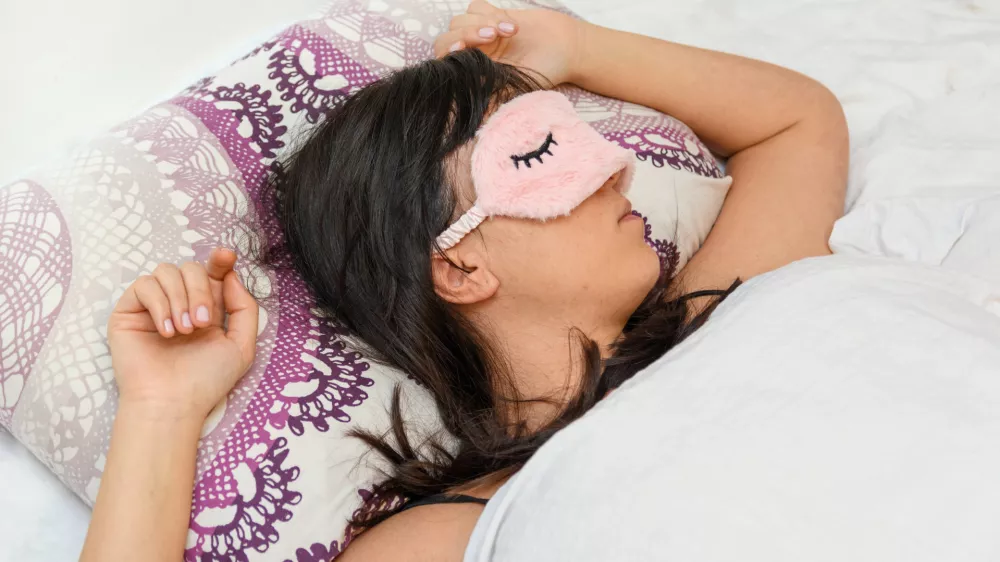 A shallow focus closeup of a female wearing a sleeping eye cover while lying on the bed / Foto: Wirestock