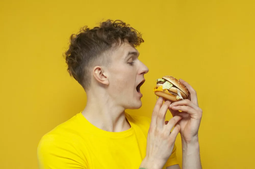 young curly guy holding a burger with his mouth open on a yellow background, a man eats fast food, close-up / Foto: Bogdan Malizkiy