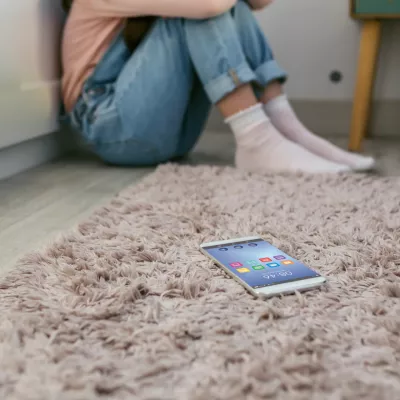 Mobile phone lying on the floor with unrecognizable sad little girl sitting behind. Selective focus on mobile in foreground