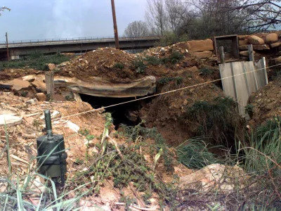 A medium close up view of a Former Yugoslavia stake mounted IMP anti-personnel mine placed near a damaged section of road, with a very visible trip wire.