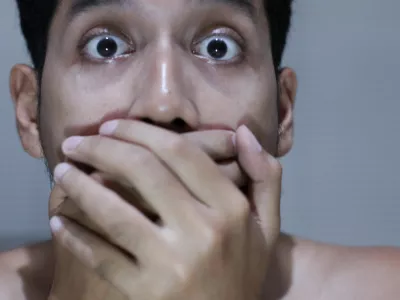 frightened asian man covering his mouth with both hands, expressing surprise, fear, or disbelief on grey blurry background / Foto: Muhammad Faris