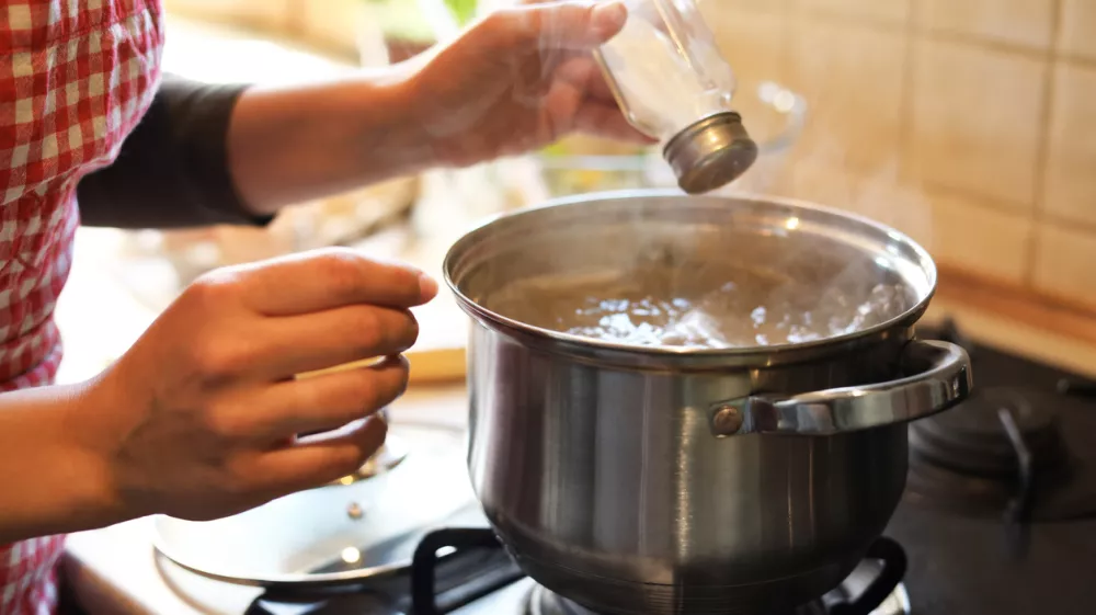 Woman adding salt to boiling water in pot on stove indoors, closeup
