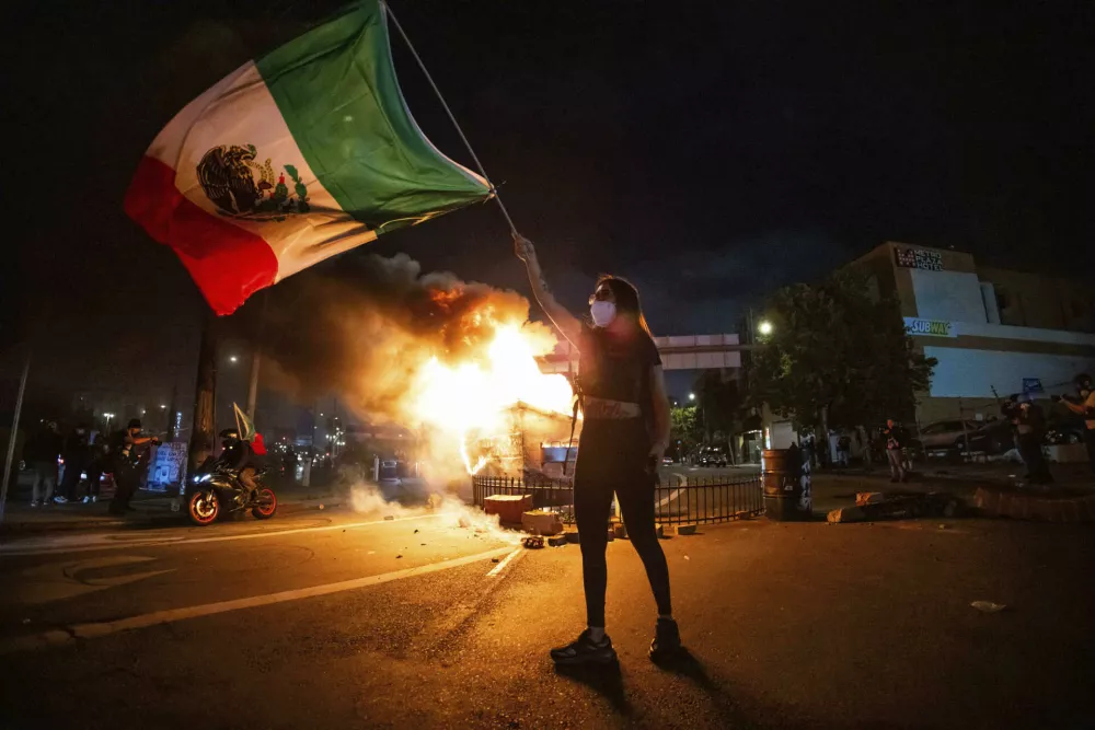 A woman waves the Mexican flag as flames erupt from a burning dumpster during a protest in downtown Los Angeles, Sunday, June 8, 2025. (AP Photo/Ethan Swope)