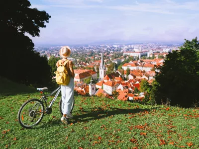 Travel Slovenia Europe. Woman tourist looking at panoramic view of cityscape with red roofs of Ljubljana from City Castle. Young girl with bicycle on top of green hill explores sights and local living / Foto: Nataliaderiabina
