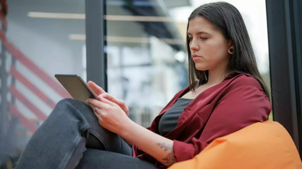 A woman sitting on a beang bag. Her eyes fixed on the screen of her tablet computer. The naturally-lit room surrounds her, with a bookshelf and window visible in the background, creating a sense of comfort and tranquility. / Foto: Bor Kankaras