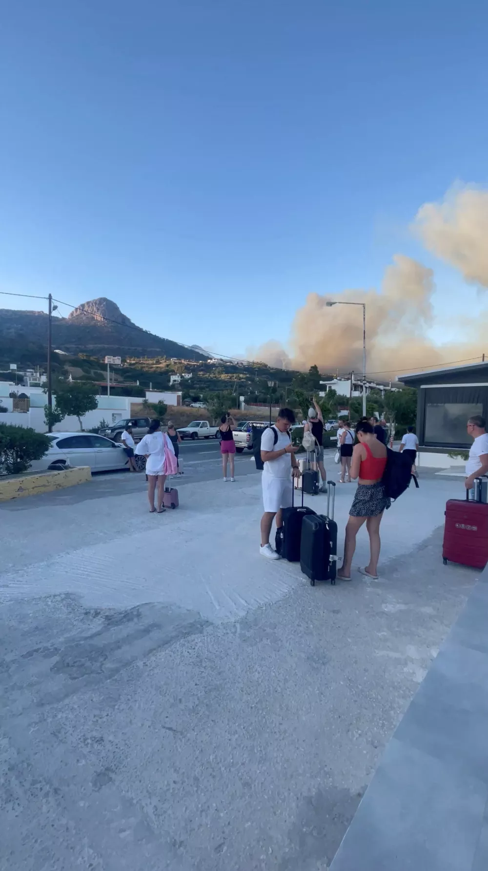 People stand with their luggage as smoke billows from a wildfire, in Ferma, on the island of Crete, Greece, July 2, 2025 in this still image from video obtained from social media. "@d_morice" via Instagram/via REUTERS THIS IMAGE HAS BEEN SUPPLIED BY A THIRD PARTY. MANDATORY CREDIT. NO RESALES. NO ARCHIVES.