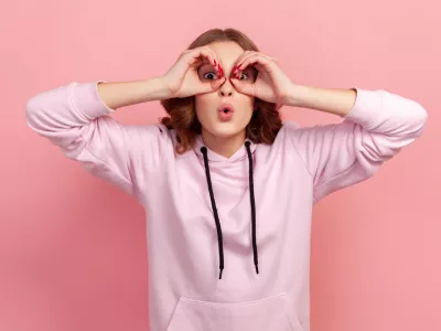 Portrait of amazed teen girl with curly hair in hoodie looking through binoculars gesture and expressing surprise, zooming vision, exploring distance. Indoor studio shot isolated on pink background / Foto: Khosrork