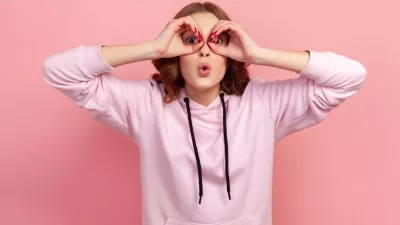 Portrait of amazed teen girl with curly hair in hoodie looking through binoculars gesture and expressing surprise, zooming vision, exploring distance. Indoor studio shot isolated on pink background / Foto: Khosrork