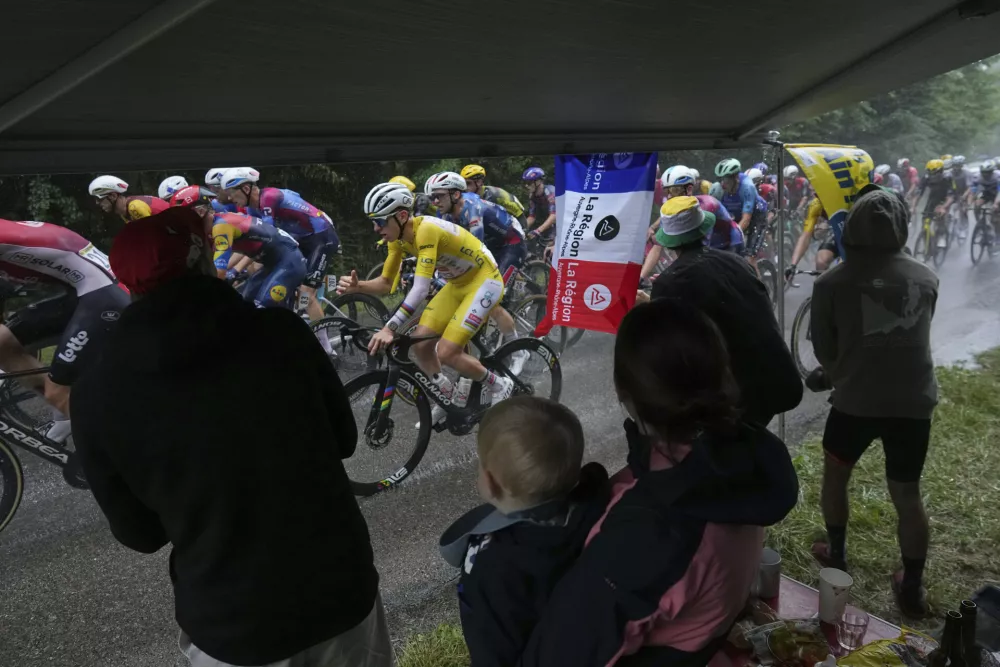 Spectators take cover under an awning as the pack with Slovenia's Tadej Pogacar, wearing the overall leader's yellow jersey, rides in the rain during the twentieth stage of the Tour de France cycling race over 184.2 kilometers (114.5 miles) with start in Nantua and finish in Pontarlier, France, Saturday, July 26, 2025. (AP Photo/Thibault Camus)