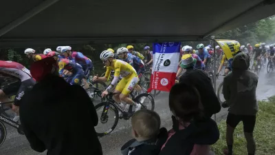 Spectators take cover under an awning as the pack with Slovenia's Tadej Pogacar, wearing the overall leader's yellow jersey, rides in the rain during the twentieth stage of the Tour de France cycling race over 184.2 kilometers (114.5 miles) with start in Nantua and finish in Pontarlier, France, Saturday, July 26, 2025. (AP Photo/Thibault Camus)