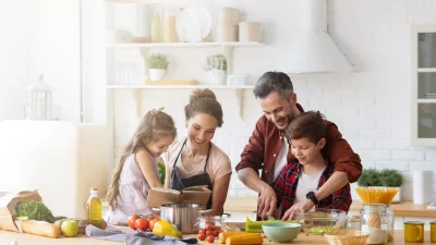 Happy family cooking together on kitchen. Mother and daughter reading recipe to father and son. Dad and boy chopping green vegetable leaf for salad. Home recreation and food preparation on weekend