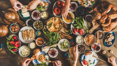 Turkish breakfast table. Flat-lay of peoples hands taking pastries, vegetables, greens, olives, cheeses, fried eggs, spices, jams, honey, tea in copper pot and tulip glasses, wide composition