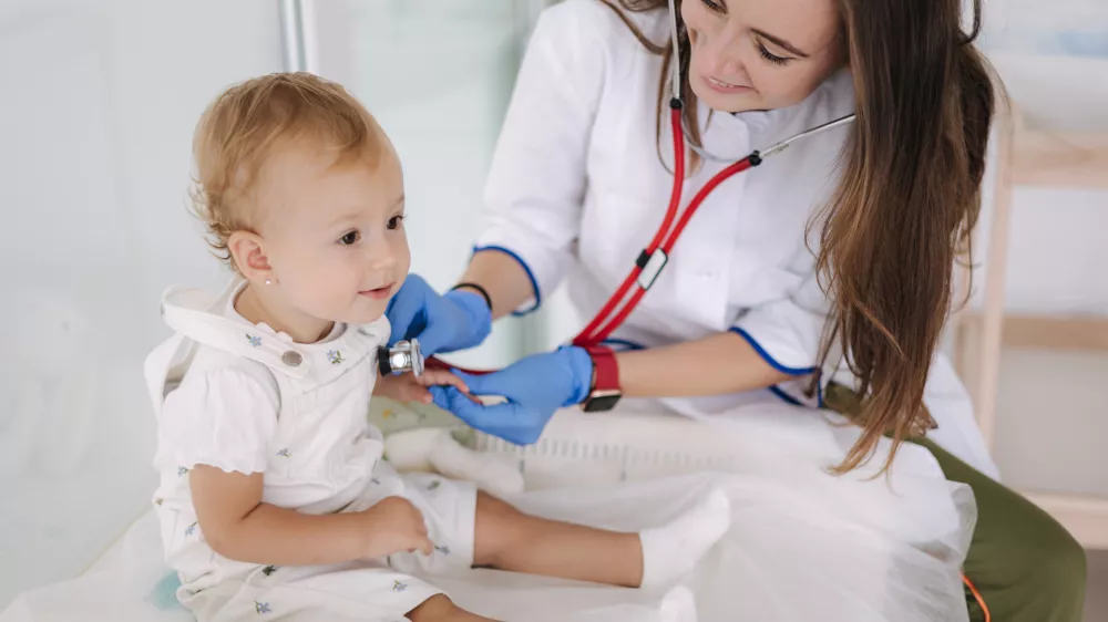 Female doctor checking child's lungs during medical checkup at the medical clinic. Woman pediatrician using stethoscope to examine breathing and heartbeat of young patient.