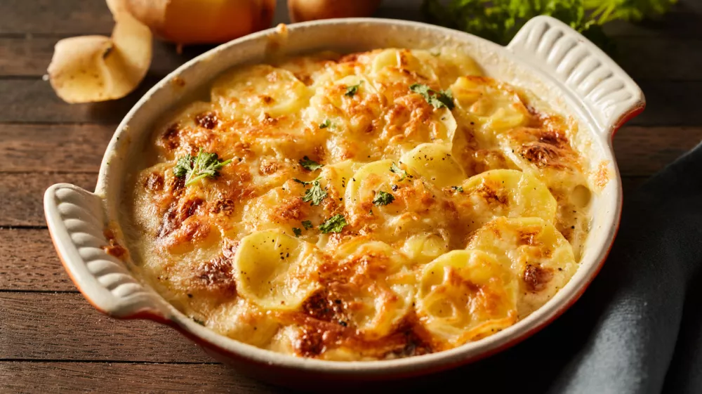 High angle of scalloped gratin potatoes topped with grated cheese and herbs in ceramic baking dish on table with towel and branches of greens / Foto: Foodandstyle, Getty