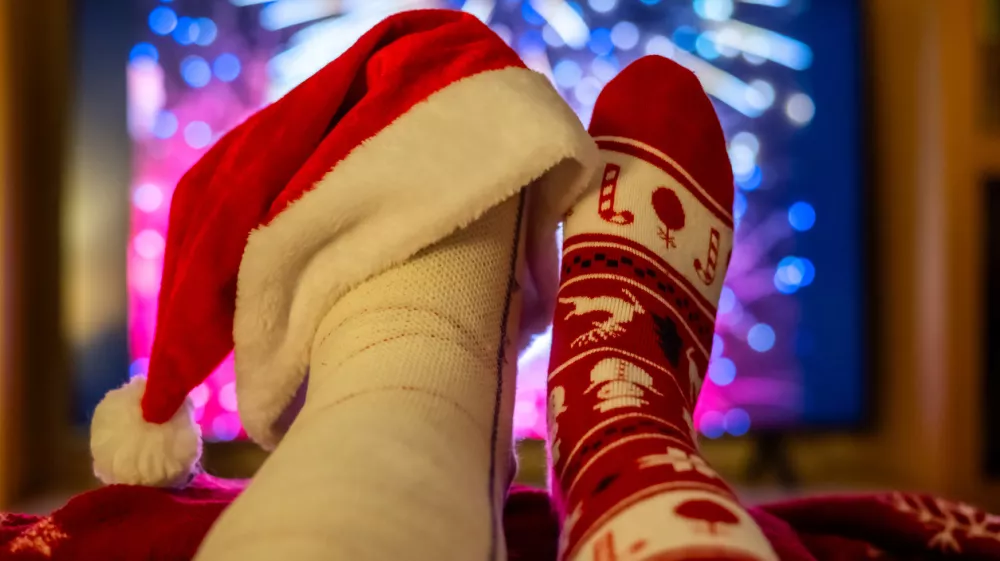Watching colorful fireworks on tv while relaxing at home, feet up and adorned with a festive christmas sock and a santa hat on a broken foot, celebrating the new year with humor and warmth / Foto: Nanci Santos, Getty Images