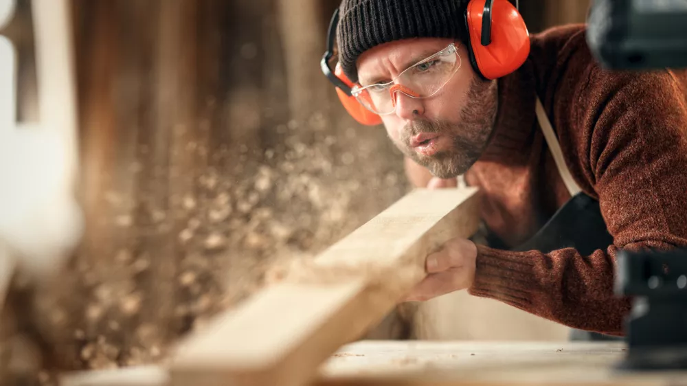Adult male woodworker in protective goggles and headphones blowing sawdust from wooden detail while working in carpentry workshop / Foto: Evgenyatamanenko