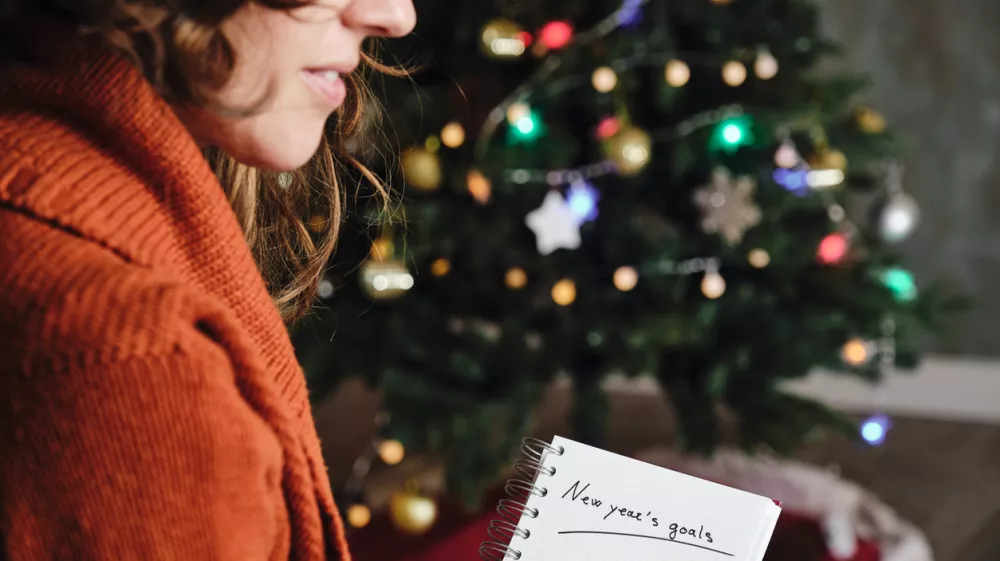 Caucasian woman smiling with orange sweater in profile with new year's goals notebook in hand with unfocused christmas tree in the background