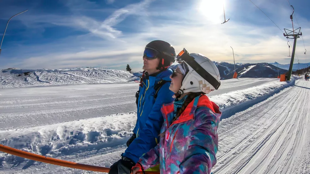 Couple going up with an anchor ski lift in Bad Kleinkirchheim, Austria. There are lot of snow caped mountains around them. They are smiling and enjoying sunny day. Winter sports. Togetherness and fun
