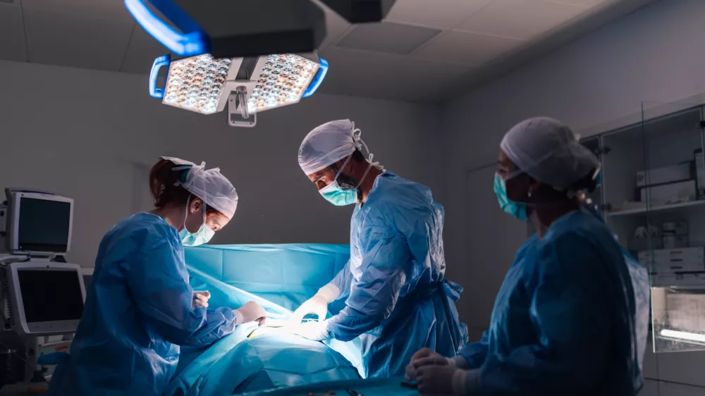 An intricate surgical procedure in progress with healthcare professionals in full surgical gear, under bright operating room lights, emphasizing the modern medical equipment and organization. / Foto: Zamrznutitonovi