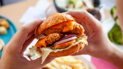 Hands holding fried chicken burger in brioche potato bun in queso sauce.