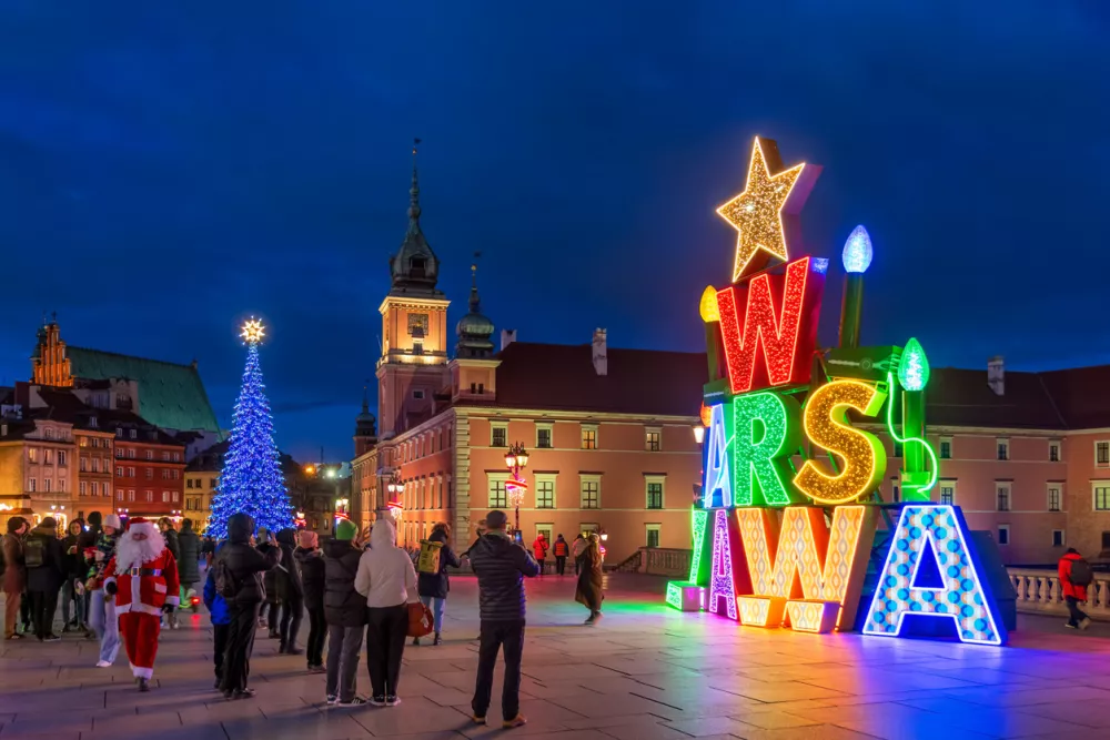 Warsaw, Poland - December 20, 2024: Illuminated Warszawa (Warsaw) sign and group of people on the Castle Square in the Old Town at night, Christmas time holiday season illumination.