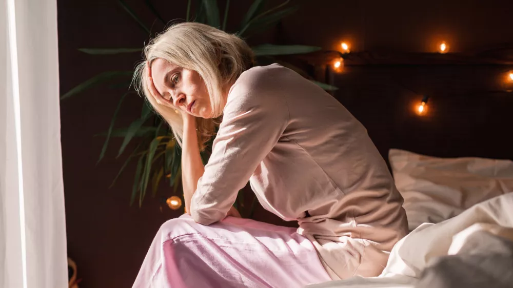 Upset middle-aged woman sit on bed in bedroom lost in thoughts thinking pondering of problem solution, depressed unhappy mature female look in distance suffer from depression, having personal problems