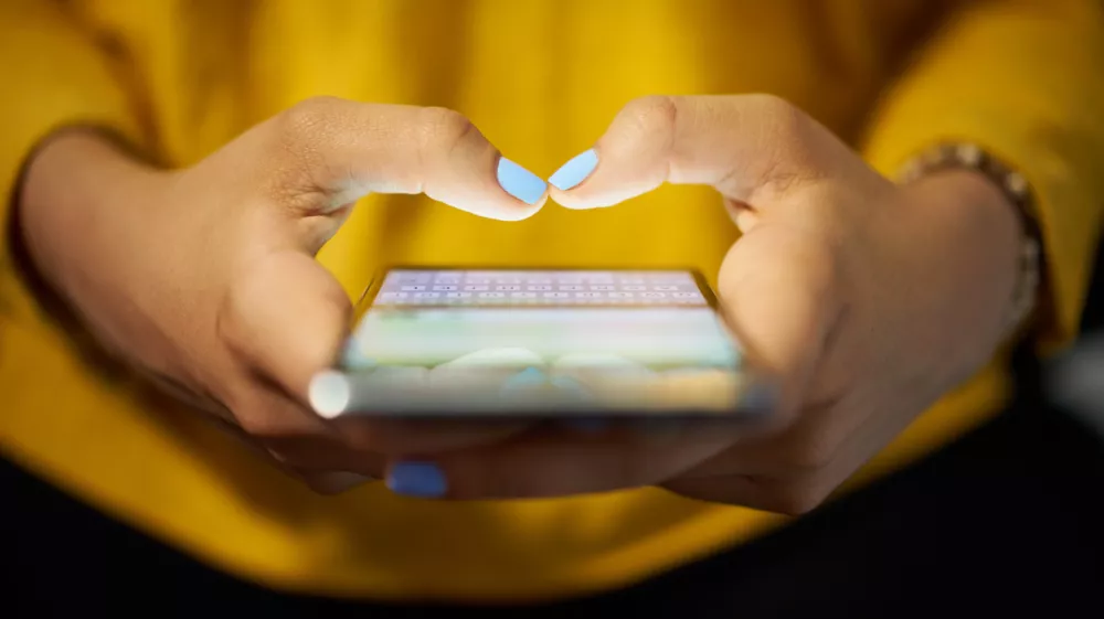 Young woman using cell phone to send text message on social network at night. Closeup of hands with computer laptop in background / Foto: Diego_cervo,getty Images/istockphoto