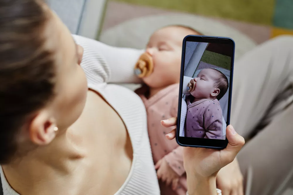Top view at unrecognizable mother carrying baby and taking photos of her adorable sleeping baby with pacifier using smartphone