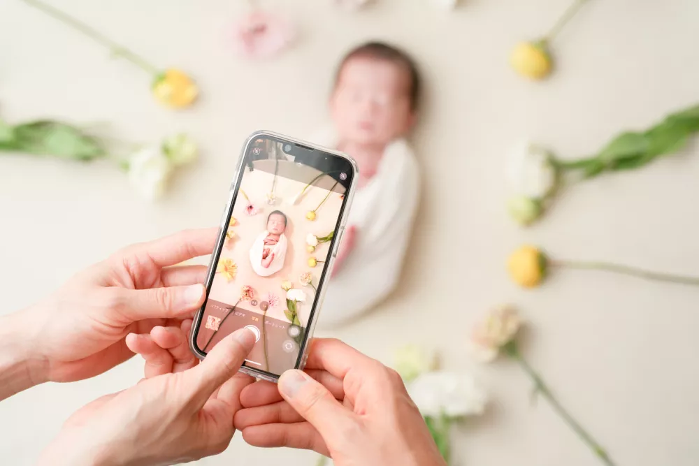 A 7-day-old Japanese newborn sleeps in a white swaddle, surrounded by spring flowers on a white background. The parents take a photo with a smartphone to save this gentle memory of new life and love.