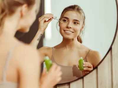 Beautiful young woman holding green bottle applying serum to face, looking in mirror, standing in bathroom at home. Concept of skin care, cosmetic procedure, moisturizing