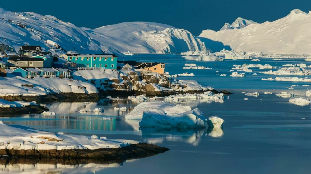 Colorful houses line the shores of a serene bay in the Arctic, surrounded by icebergs and snow-covered mountains, reflecting the warm glow of the sunset on the water.