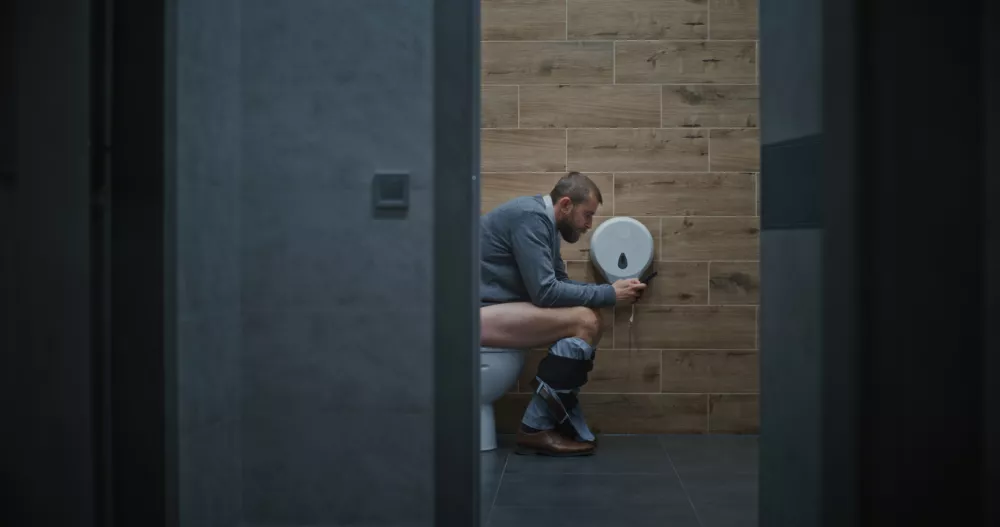 Businessman Using Smartphone, Sitting on the Toilet. Man Scrolling Social Networks and Media Content, Feeling Relaxed in Restroom in Modern Office During Work. Lifestyle and Daily Routine. Full Shot.