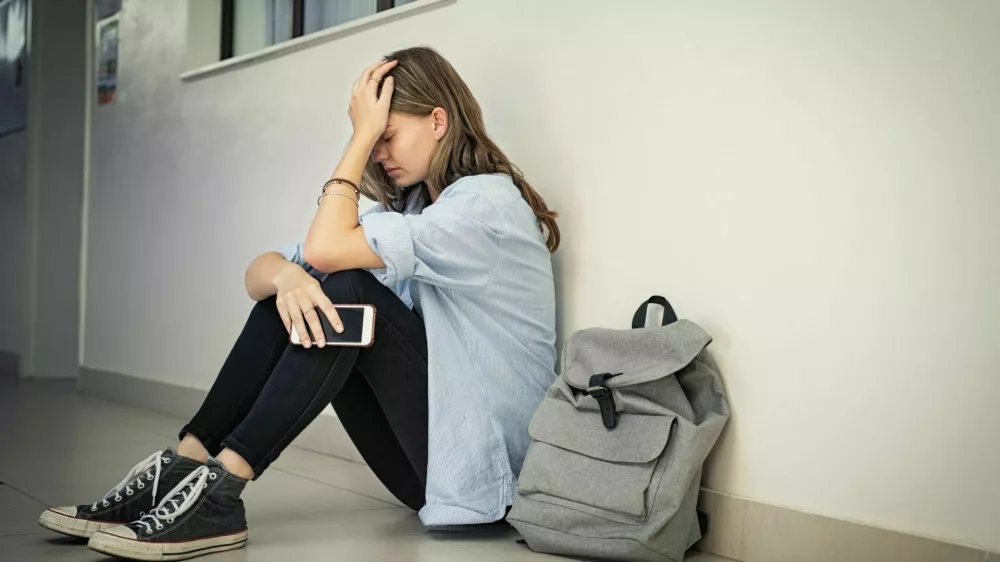 Upset and depressed girl holding smartphone sitting on college campus floor holding head. University sad student suffering from depression sitting on floor at high school. Lonely bullied teen in difficulty with copy space. / Foto: Ridofranz