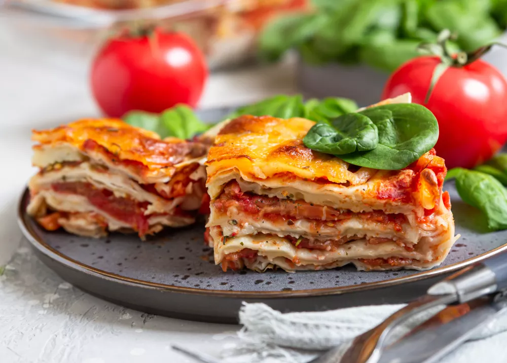 Italian lasagna with tomato sauce and cheese served with tomatoes and spinach, light concrete background. Homemade vegetarian lasagna. Selective focus. / Foto: Porosolka