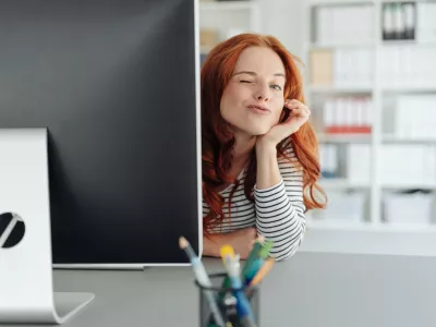 Playful young businesswoman winking at the camera as she peers around the edge of a large desktop monitor in a spacious office