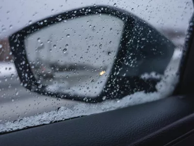 Raindrops cover the side mirror of a car while driving on a road in winter. Snow gathers on the car door with gray skies above in the background.