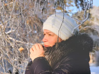 Woman in the blue padded jacket and white cap under snowfall at the street. Pensive attractive mature woman with a quiet smile. Sunny winter vacation outdoors. Deep breathing freshness